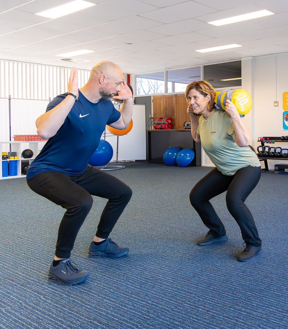 Trainer begeleidt een deelnemer met een sandbag tijdens een oefening in de studio