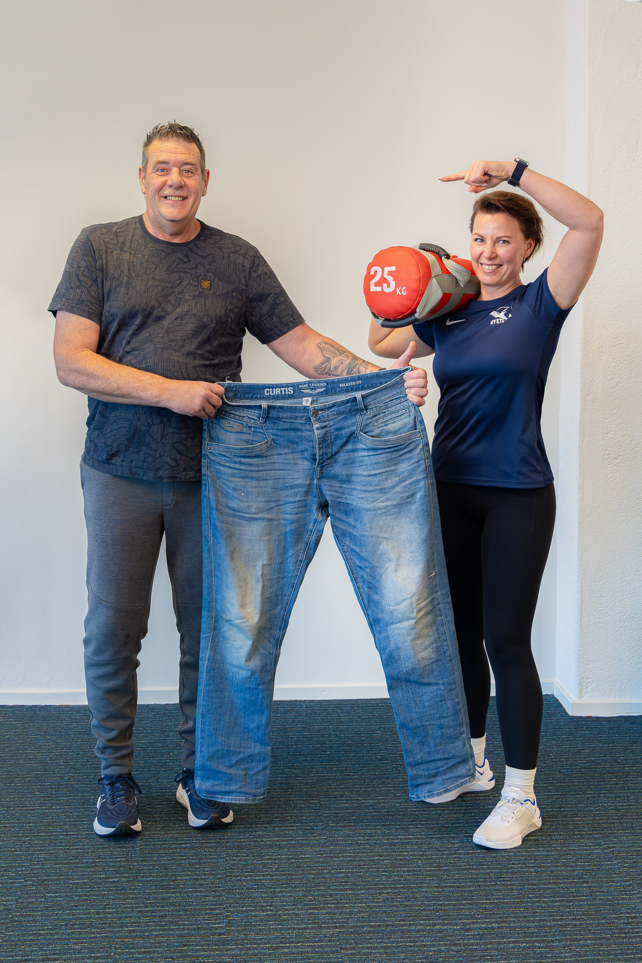 René met zijn oude spijkerbroek na 25 kilo afvallen, naast zijn trainer