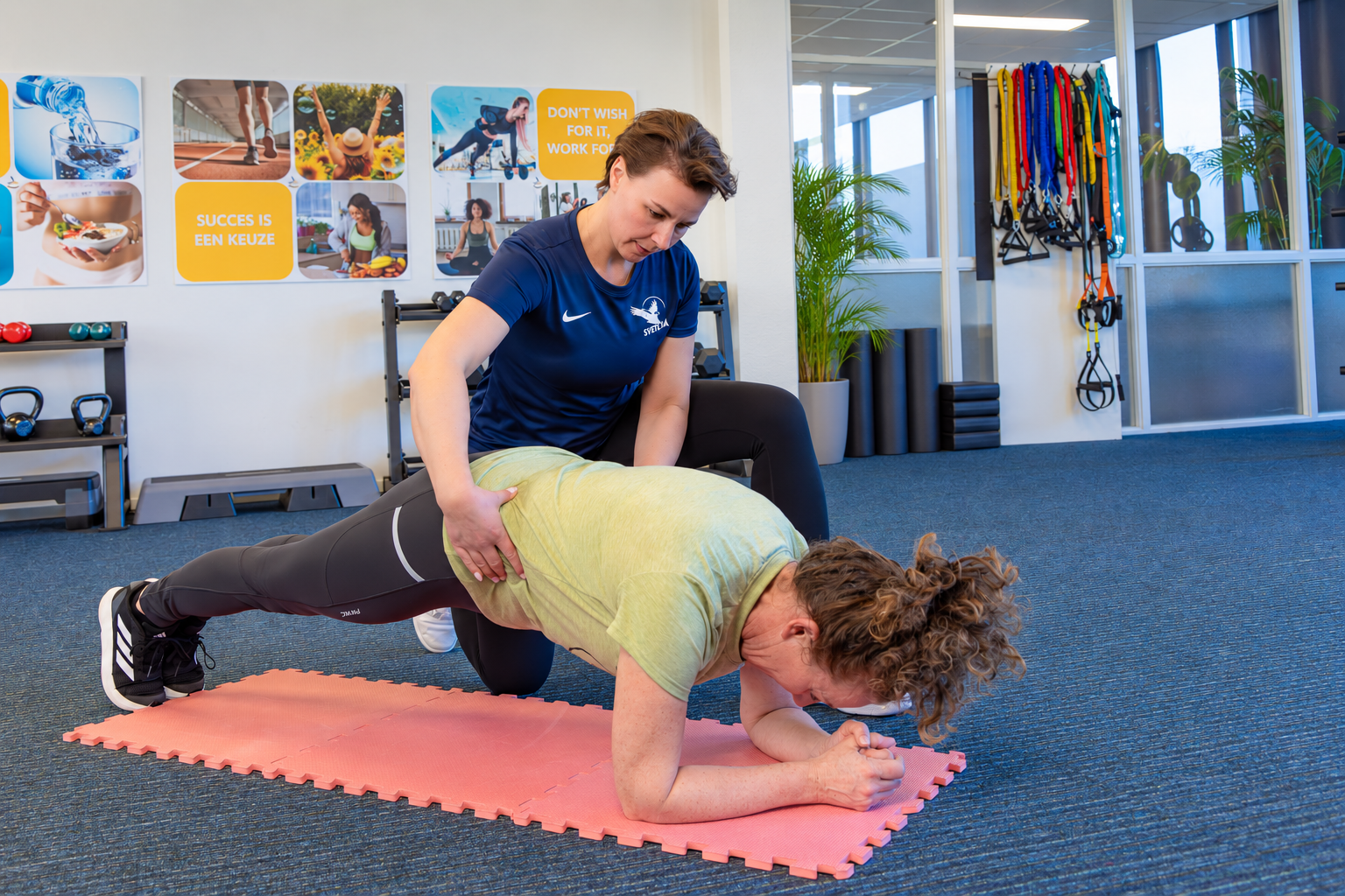 Trainer begeleidt een deelnemer tijdens een oefening op een mat in de studio